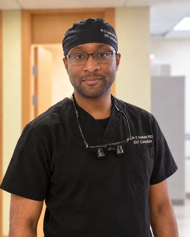 Mr Eugene Omakobia smiling in black surgical scrubs and glasses, with surgical loupes