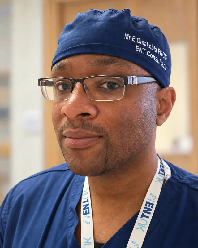 Mr Eugene Omakobia smiling in navy surgical scrubs, cap and glasses